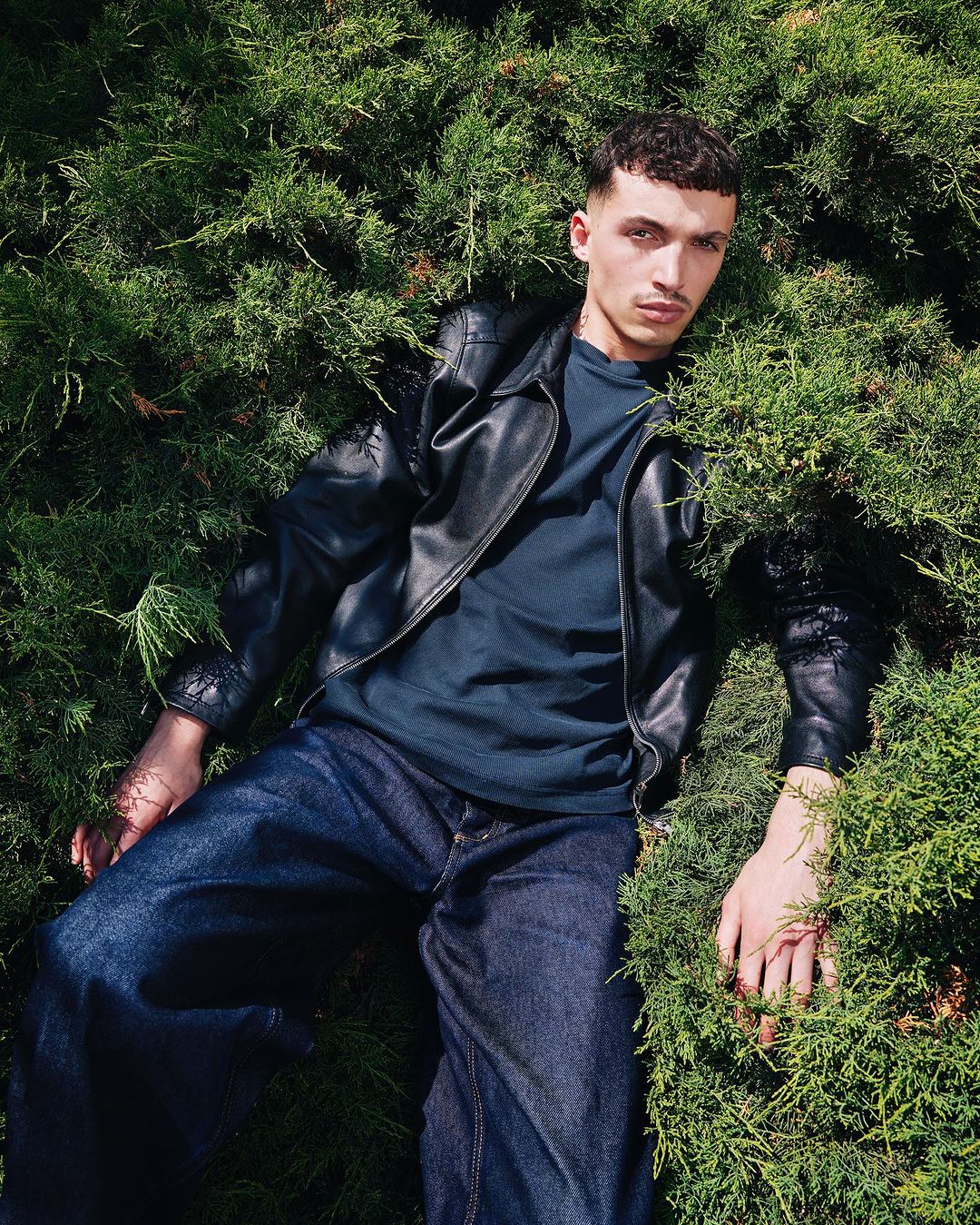 Young man in a black leather jacket and jeans lies among green bushes, looking at the camera with a neutral expression.