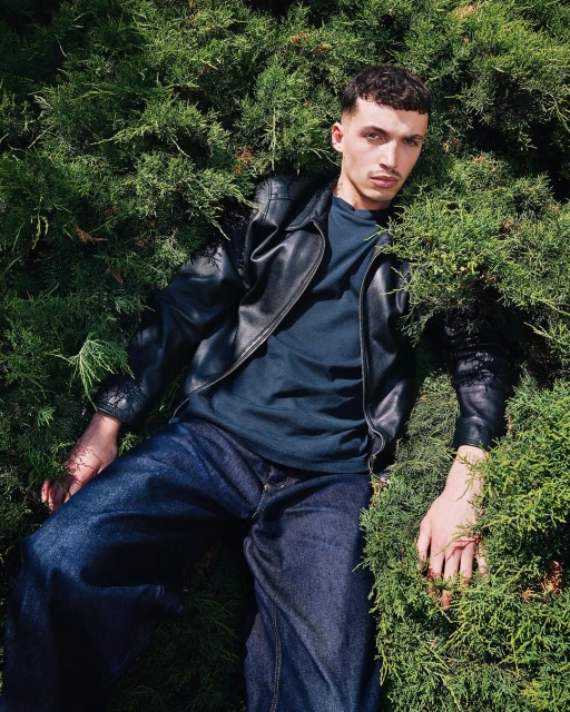 Young man in a black leather jacket and jeans lies among green bushes, looking at the camera with a neutral expression.