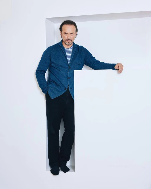 Portrait of a middle-aged man in a blue jacket and black pants, standing in a white doorway against a white background in a photo studio.