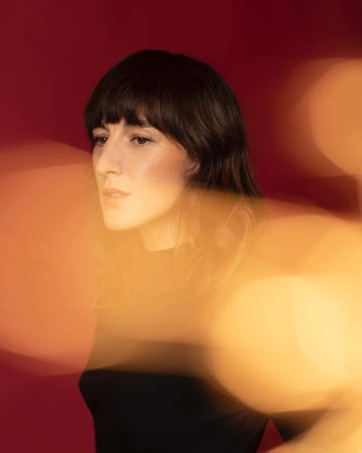 Portrait of a young woman with dark hair, shot in front of a dark red backdrop with gold bokeh lights creating a diffused effect.