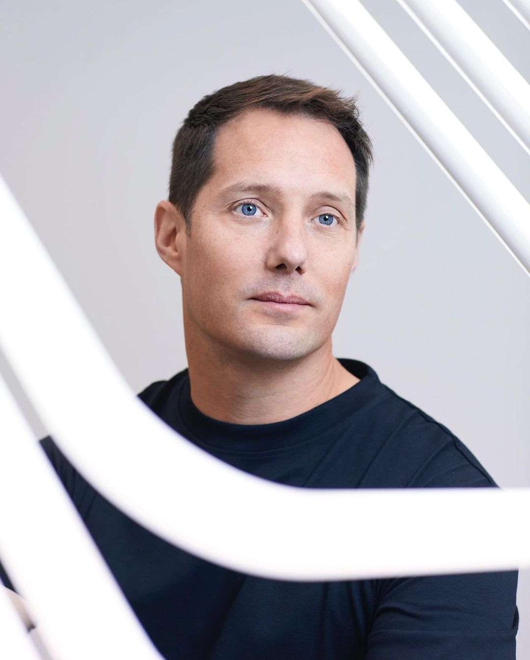 Close-up portrait of an adult male with blue eyes wearing a dark shirt, framed by white architectural elements in a brightly lit studio.
