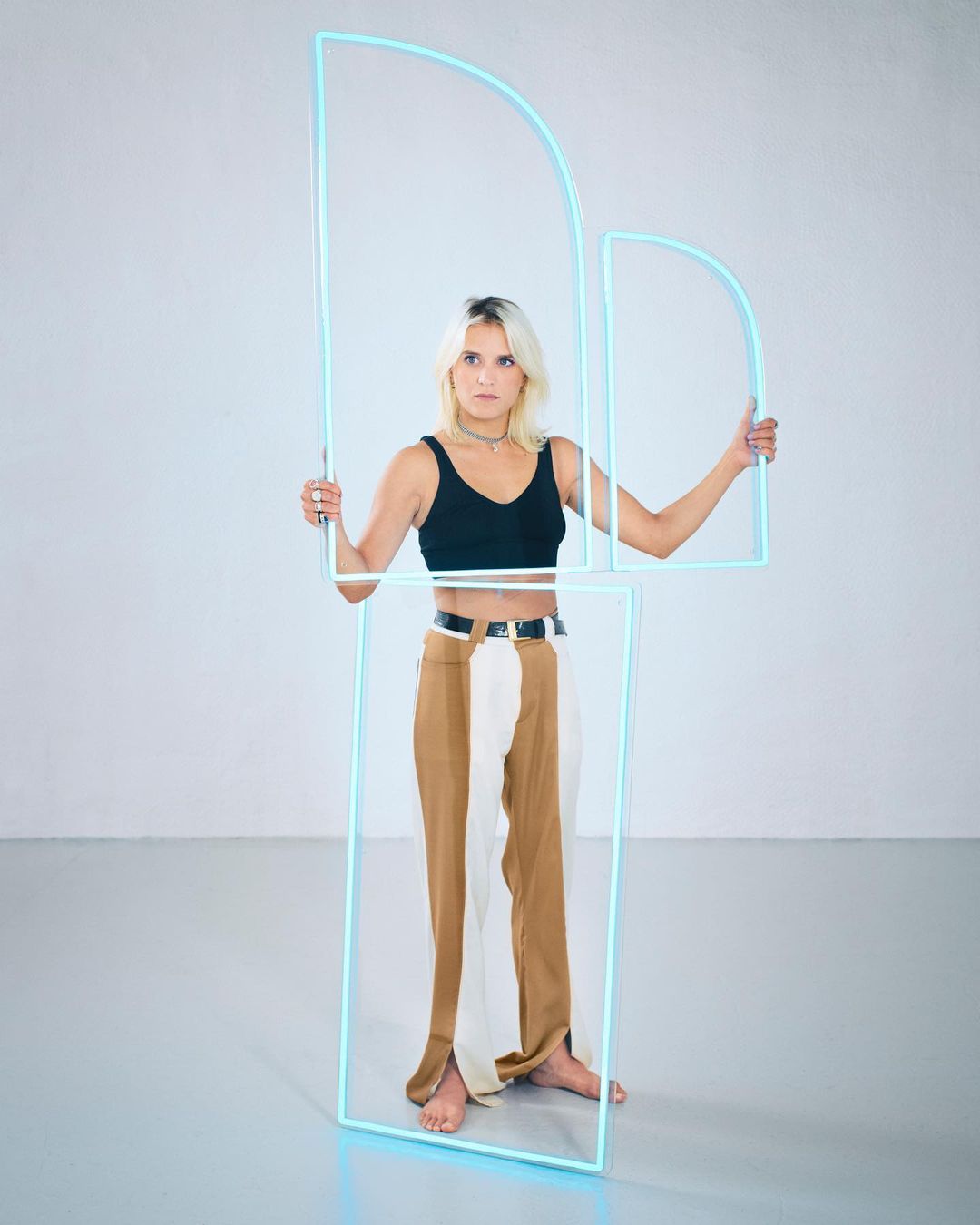 Full-length shot of a barefoot woman holding geometric neon light tubes in a studio, wearing a black top and two-tone pants.