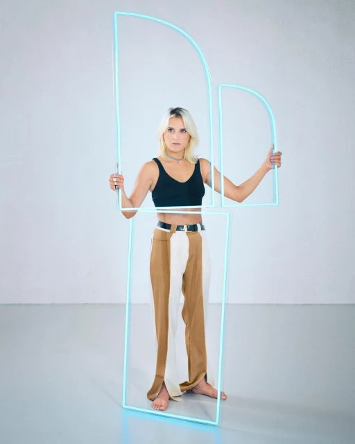 Full-length shot of a barefoot woman holding geometric neon light tubes in a studio, wearing a black top and two-tone pants.