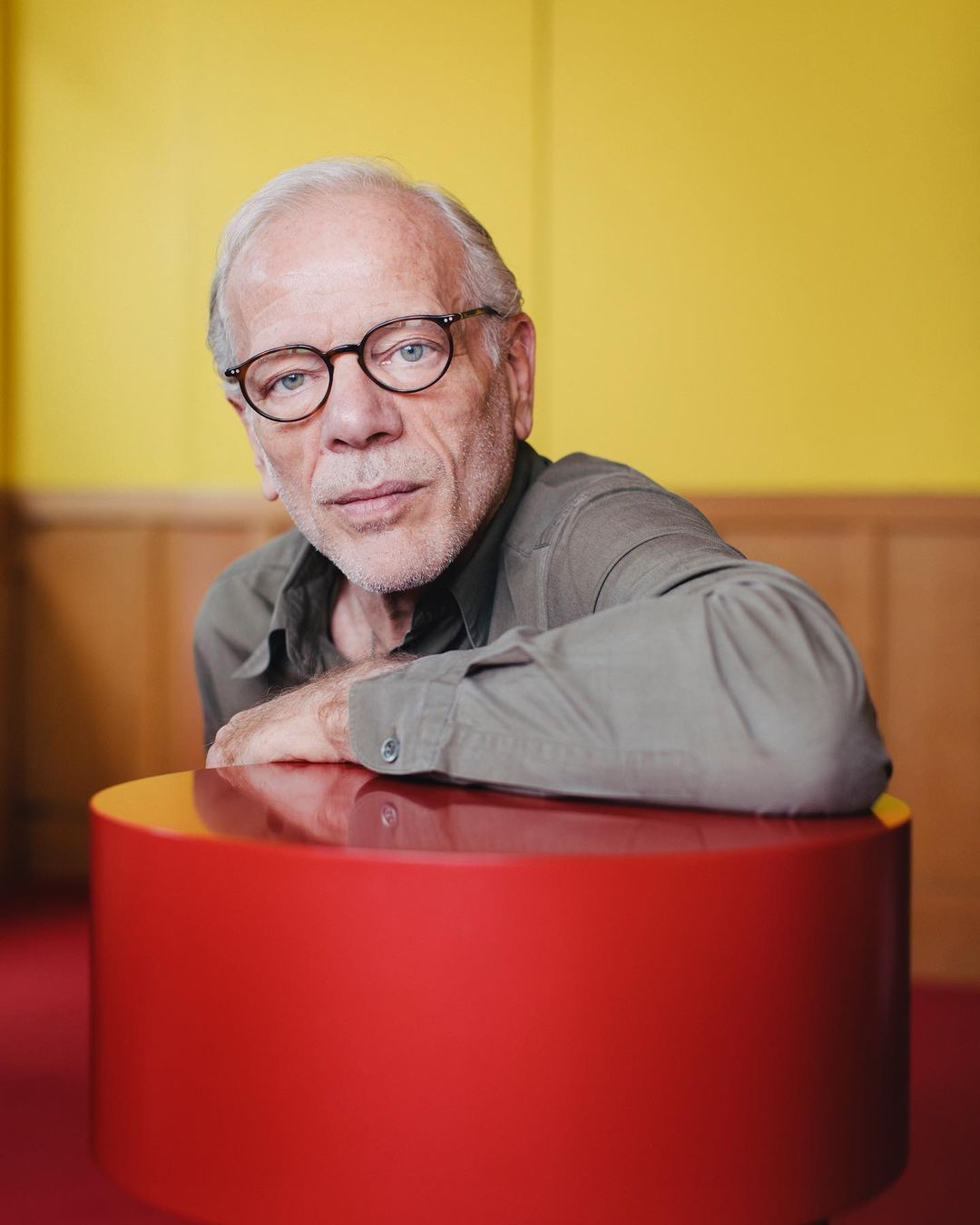 Portrait of an elderly man with glasses resting his hands on a round red table in a yellow and brown indoor setting.