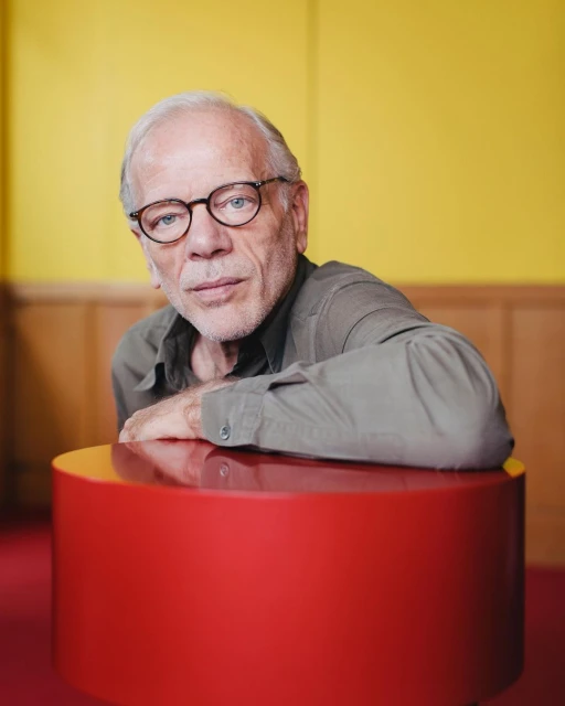 Portrait of an elderly man with glasses resting his hands on a round red table in a yellow and brown indoor setting.