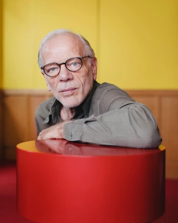 Portrait of an elderly man with glasses resting his hands on a round red table in a yellow and brown indoor setting.