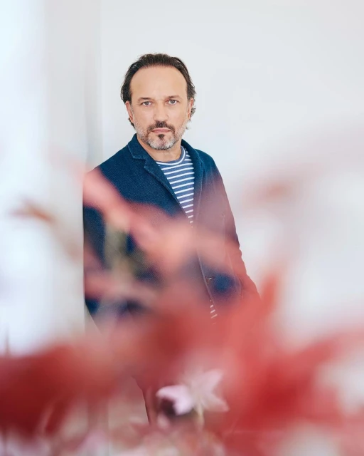Portrait of a middle-aged man with dark hair and a beard, wearing a blue jacket and striped shirt, posing indoors with a soft background.