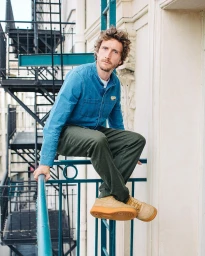 Man in a blue denim shirt and dark trousers crouching on a turquoise balcony railing in Paris.
