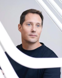 Portrait of a white man with short dark hair and blue eyes wearing a dark t-shirt, framed by white structural elements in Paris