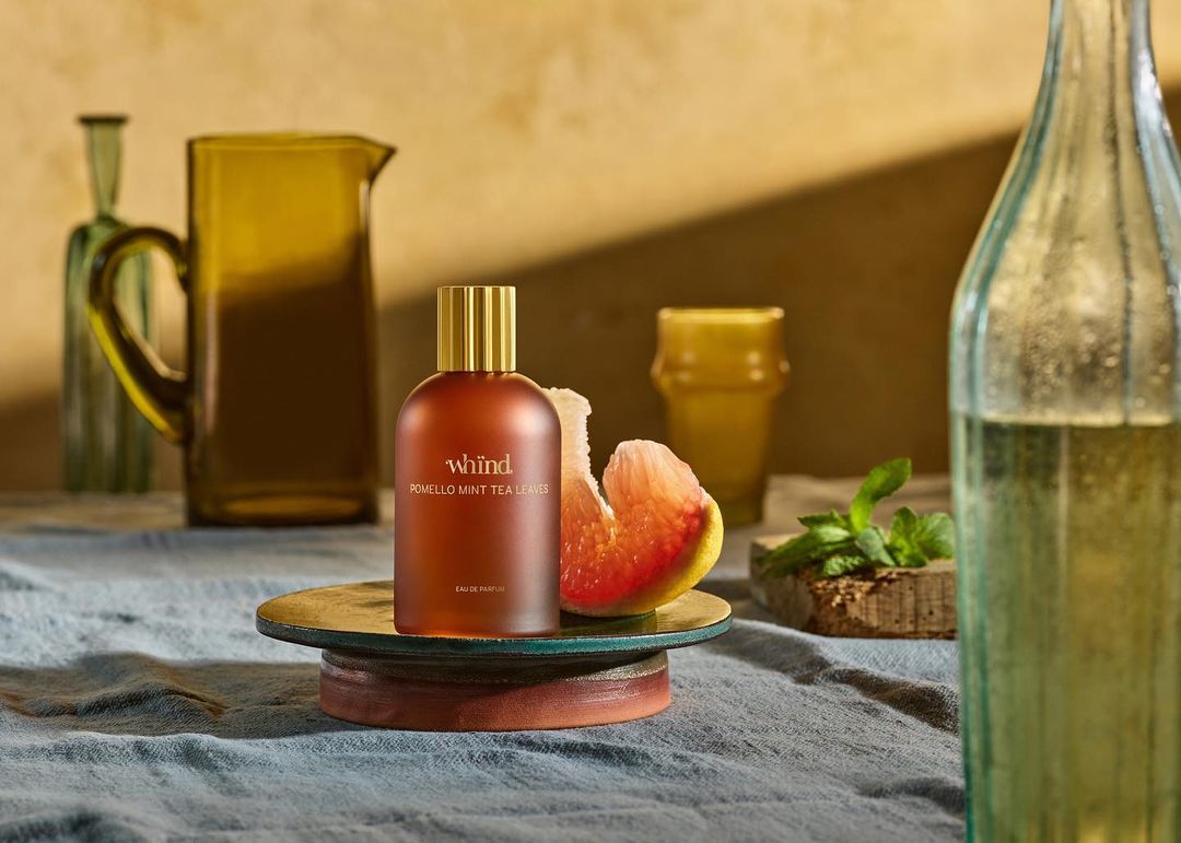 Still life shot of a brown perfume bottle with a gold cap displayed alongside a grapefruit slice and mint leaves on a blue tablecloth.