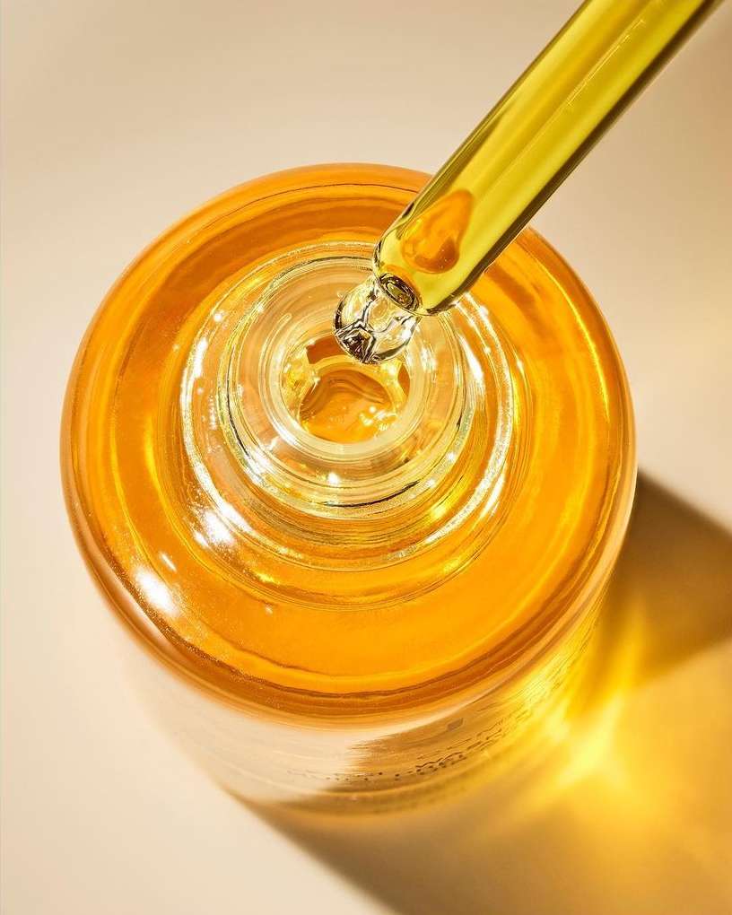 Close-up of a glass bottle with yellow liquid and a dropper; the bottle is on a beige surface and casts a soft shadow.