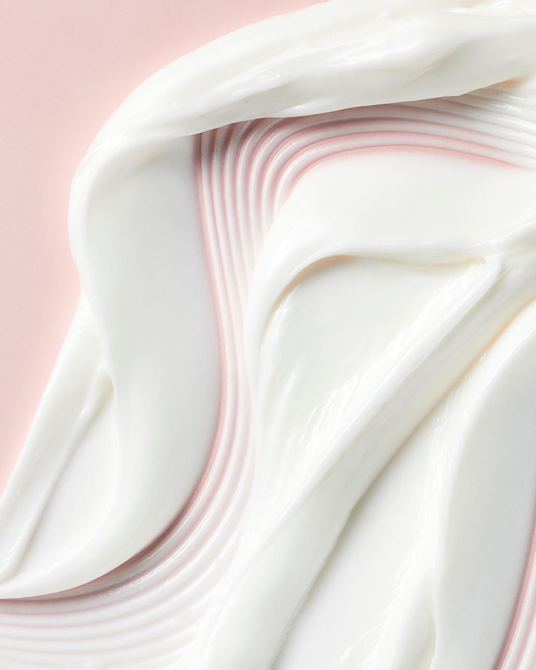Close-up of thick, creamy white cosmetic product swirled on a light pink background, showcasing smooth texture and gentle ridges.