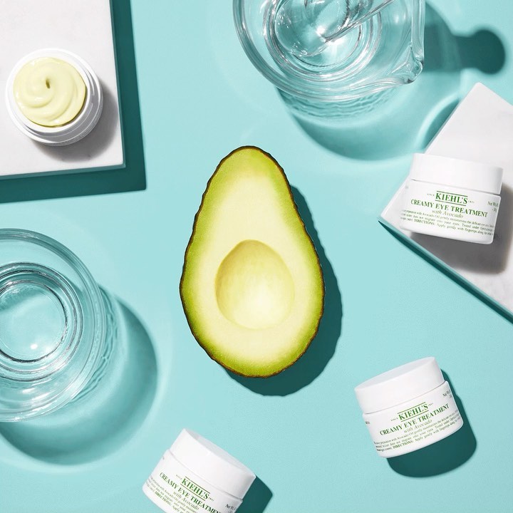 Overhead shot of avocado half, eye cream jars, and glass containers on a light blue background, ideal for beauty product promotion.