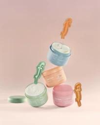 Four pastel cosmetic jars stacked diagonally with floating letter f shaped product samples against a pink backdrop