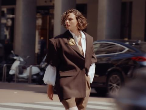 Stylish young adult female model poses in a brown blazer and white shirt on a city crosswalk, with a scooter and black car in the background.