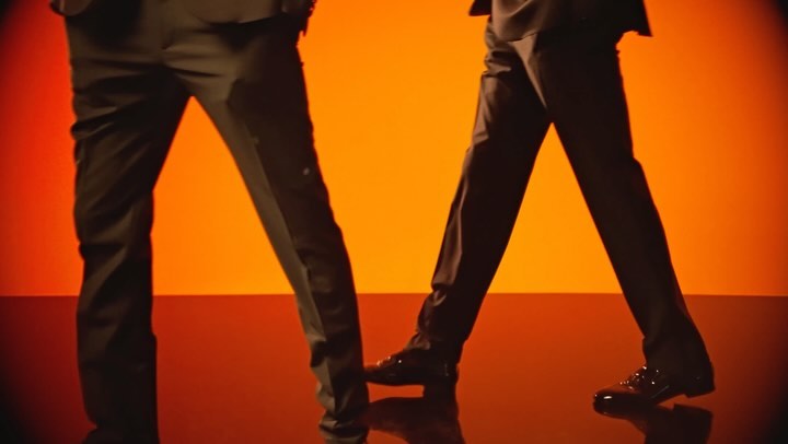Close-up of two people in dark suits walking on a reflective surface with a vivid orange background, focusing on legs and shoes.