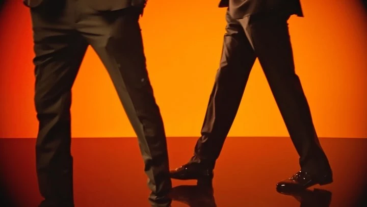 Close-up of two people in dark suits walking on a reflective surface with a vivid orange background, focusing on legs and shoes.