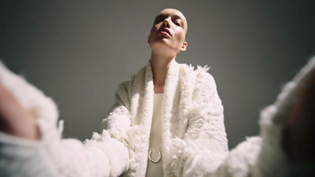 Elegant shot of a bald adult woman in a white outfit and furry coat, gazing upward in a studio setting with diffused lighting.
