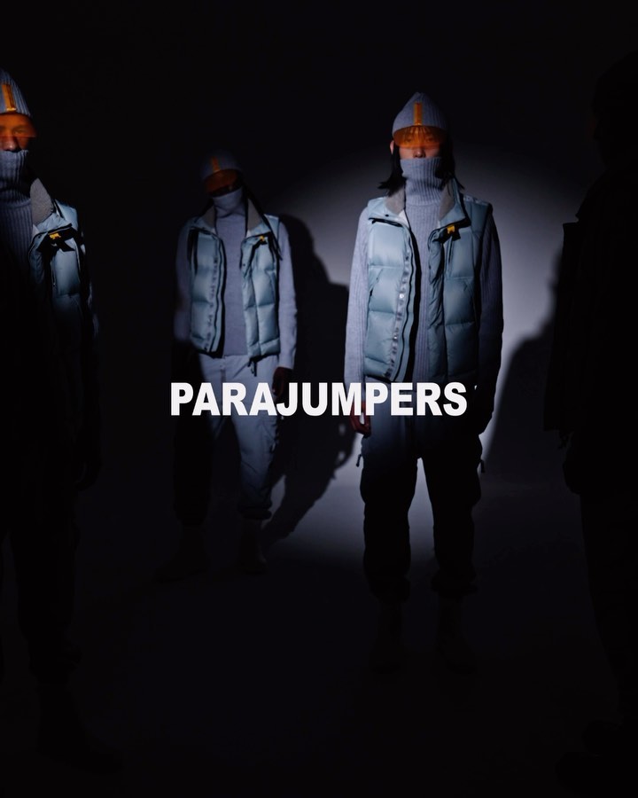 Four fashion models stand in a dimly lit studio wearing puffer vests, turtlenecks, and matching knit caps, showcasing winter outerwear.