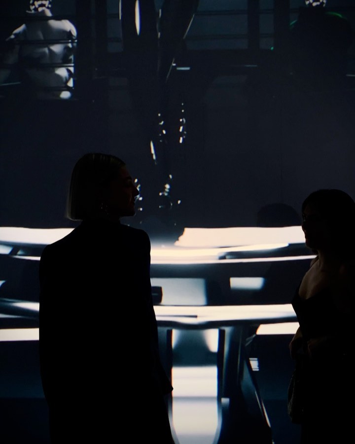 Two figures silhouetted in a dimly lit space with tables, creating an atmosphere of mystery, and an indiscernible scene in the background.