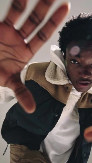 Close up shot of a young adult male with curly hair, wearing a jacket and hoodie, reaching towards the camera in a neutral indoor space.
