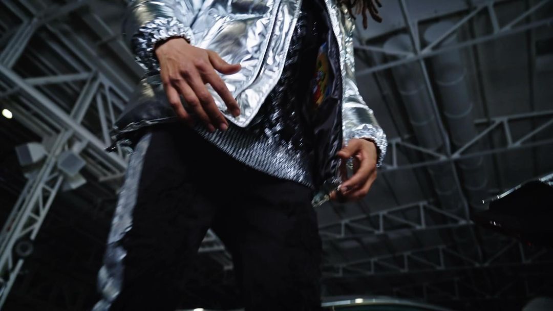 Low-angle shot of a person in a silver jacket and black pants with silver stripes, set against a dark, industrial background with visible beams.
