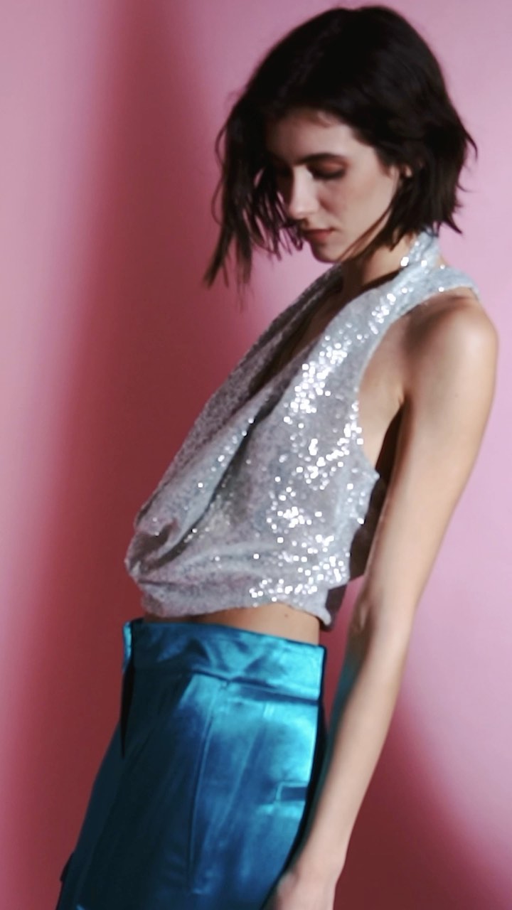 A woman in a sequin top and blue satin pants poses against a pink background in a studio, showcasing a fashion or beauty campaign.