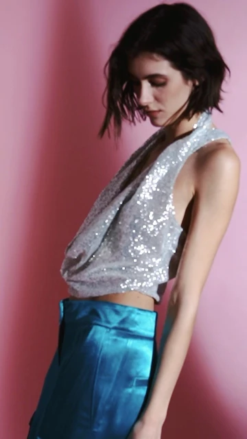 A woman in a sequin top and blue satin pants poses against a pink background in a studio, showcasing a fashion or beauty campaign.
