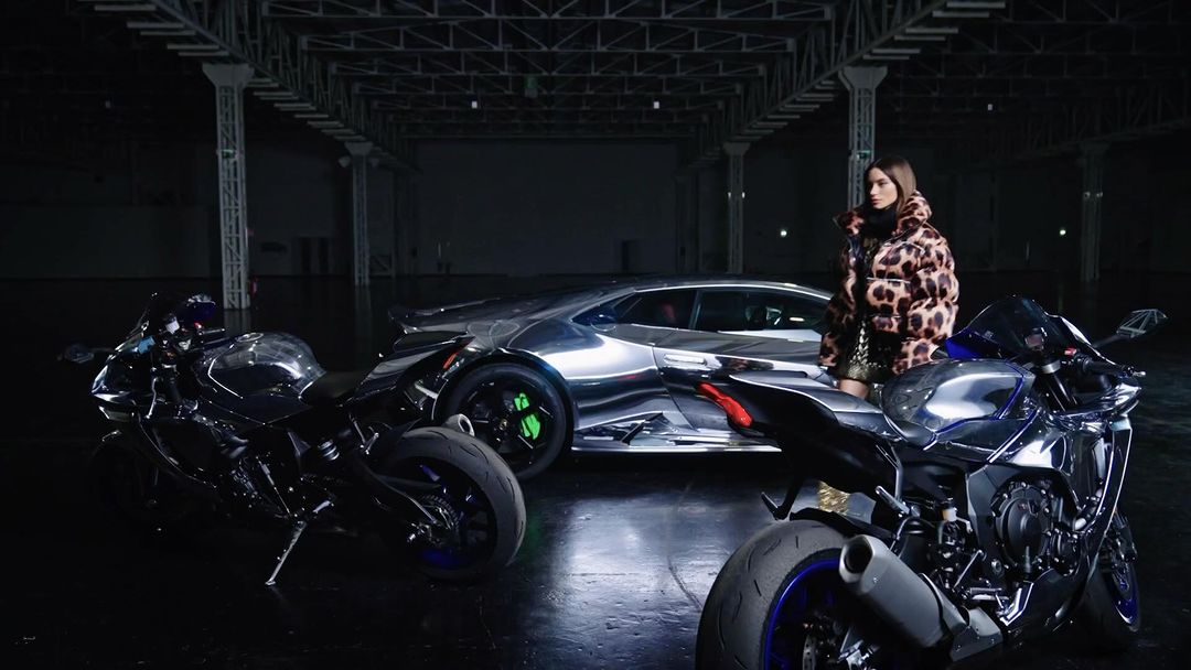 A woman poses in a leopard print coat next to a silver Lamborghini and two black motorcycles in a dark, industrial space.