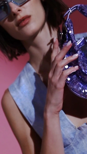 A woman with short brown hair poses with a shiny purple purse, wearing a denim dress and blue sunglasses in a studio setting.