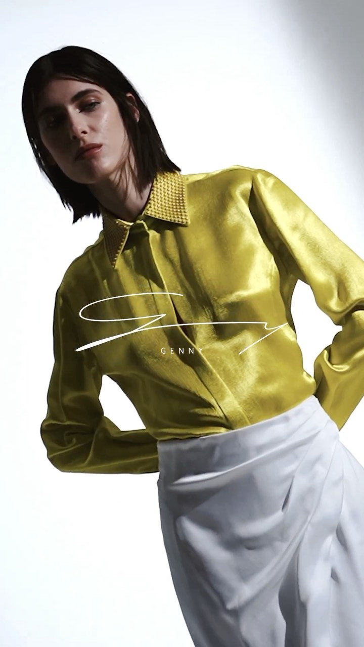 A fair-skinned woman in a yellow, long-sleeved silk blouse and white skirt poses in front of a stark white backdrop.