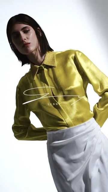A fair-skinned woman in a yellow, long-sleeved silk blouse and white skirt poses in front of a stark white backdrop.