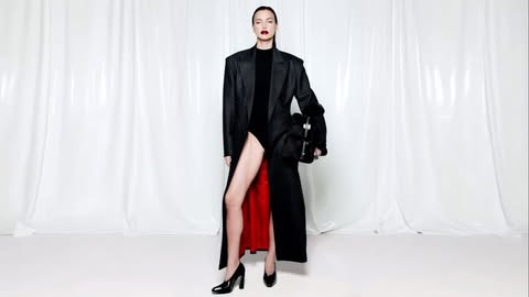 Woman in black coat, dress, and high heels poses in a photo studio in front of a white curtain backdrop, holding a black purse.
