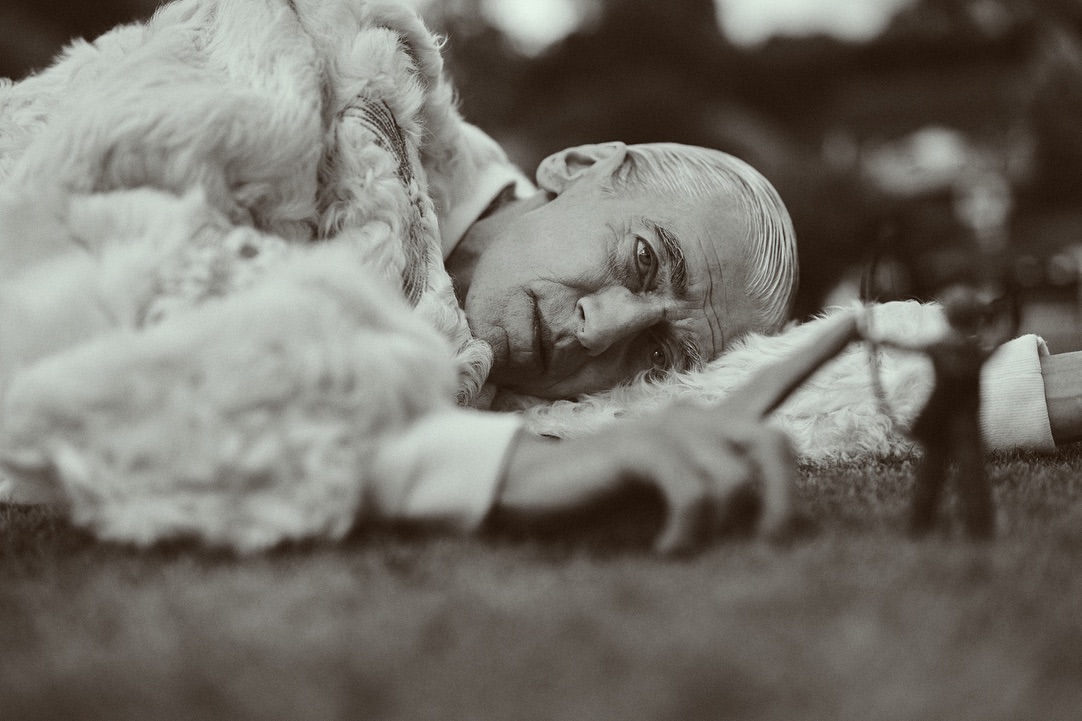 Monochrome portrait of an elderly man lying on grass wearing a fur coat, gazing directly at the camera, conveying a sense of calm.