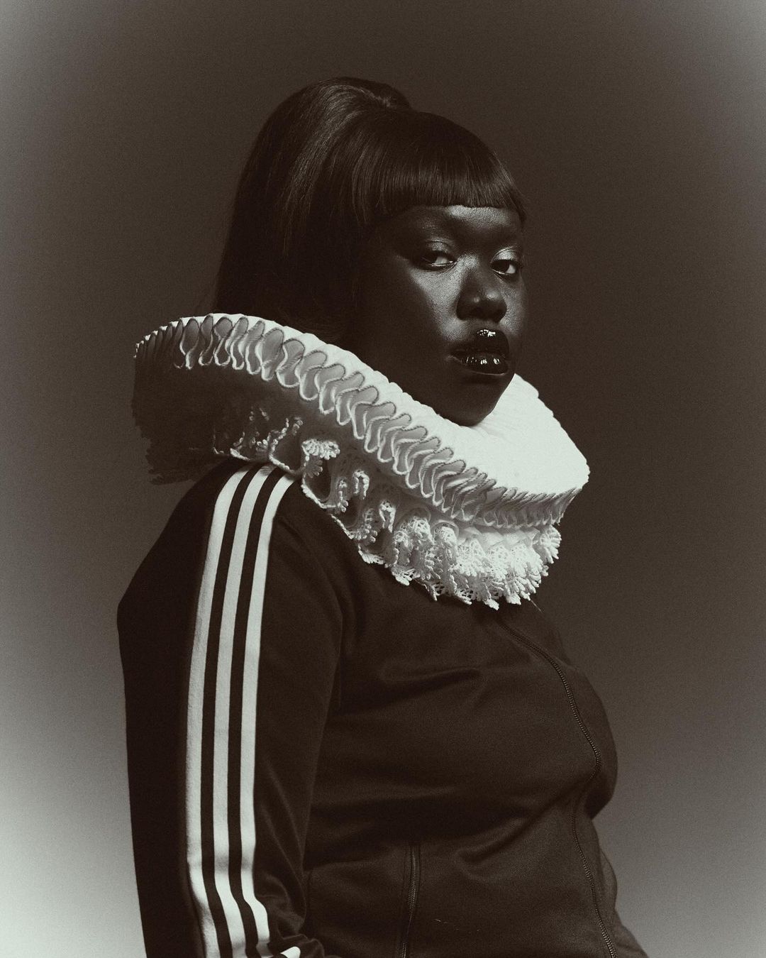 Woman with dark skin and an afro-style hairstyle wearing a tracksuit jacket and a white ruffled Elizabethan collar against a grey background.