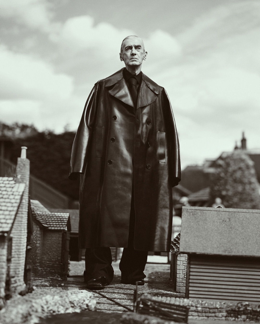 Monochrome portrait of a mature man in a black leather coat standing amidst a miniature village against a cloudy sky backdrop.
