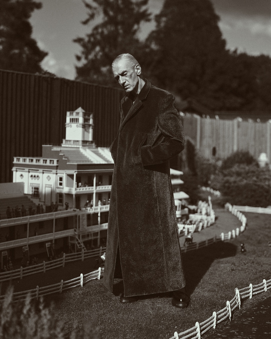 Sepia photograph of an older adult male in a long coat standing next to a miniature golf course with small-scale buildings and landscaping.