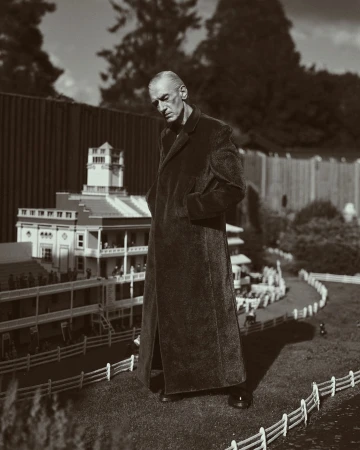 Sepia photograph of an older adult male in a long coat standing next to a miniature golf course with small-scale buildings and landscaping.
