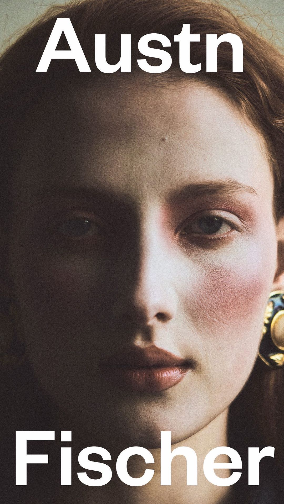 Close-up portrait of a fair-skinned young woman with soft red blush, a serious expression, and statement earrings in a dimly lit setting.