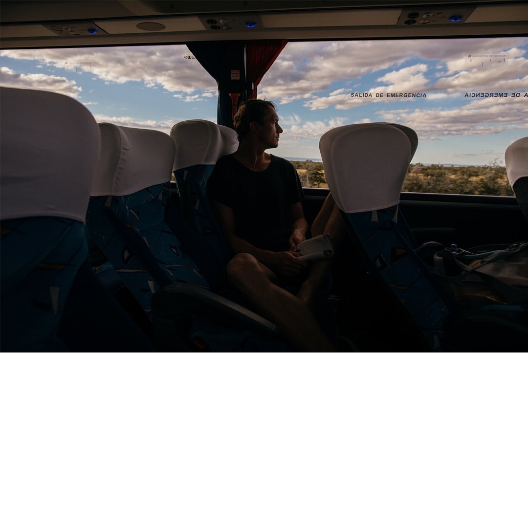 Adult sits alone on a bus, looking out the window at rural landscape under a blue, cloudy sky. Bus seats are visible around.