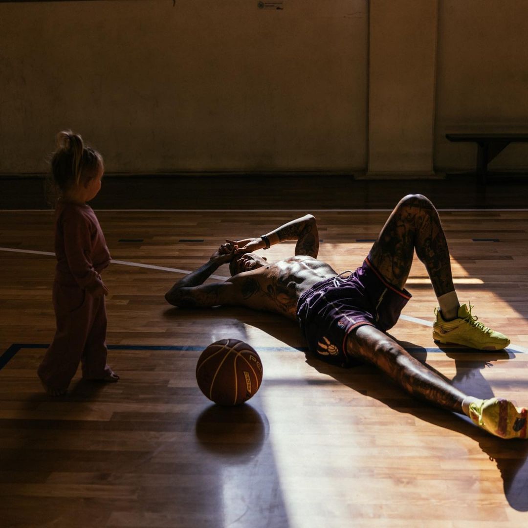 Tattooed man in athletic wear lying on basketball court with ball, looking at standing toddler near the wall in natural light.