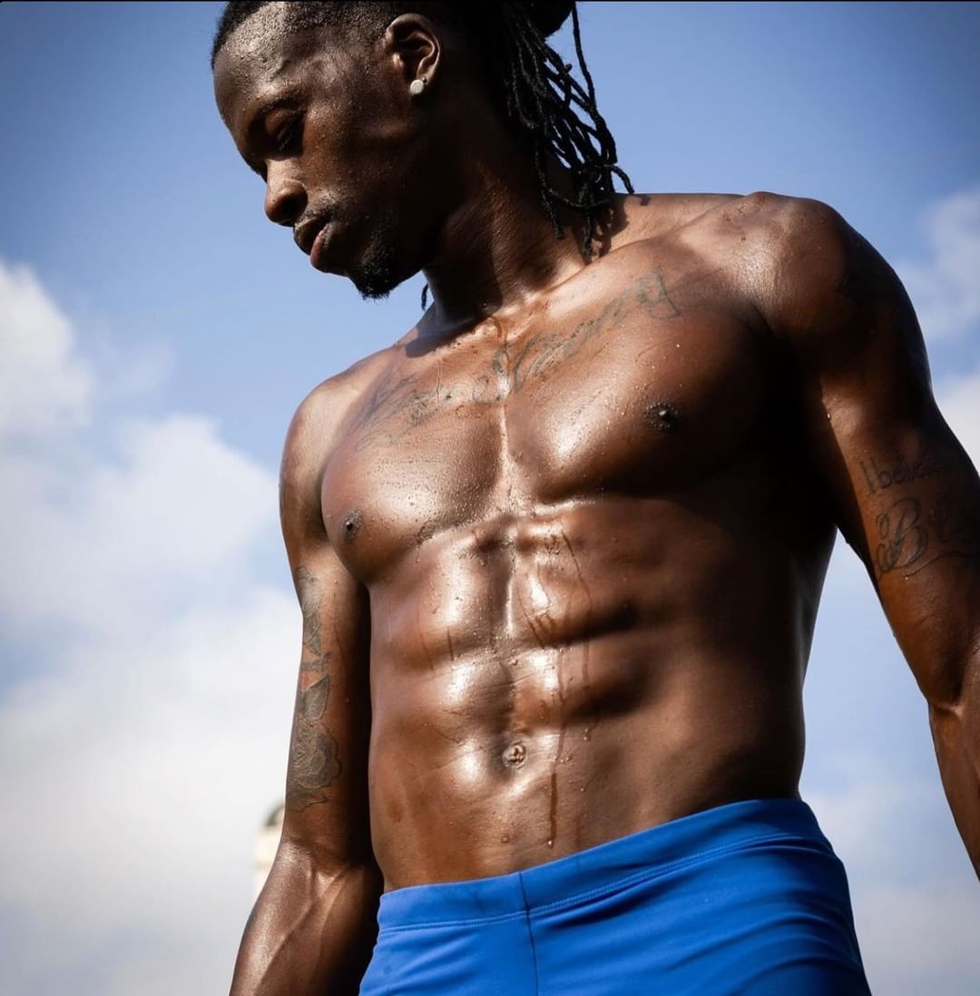 Athletic African-American man with dreadlocks shows muscular torso outdoors against a blue sky wearing blue shorts, featuring tattoos.