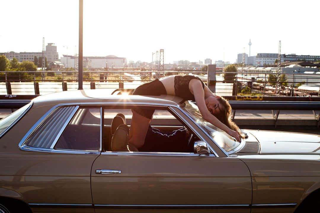 An adult woman poses on the hood of a classic tan car on a city street with buildings in the background on a sunny day.