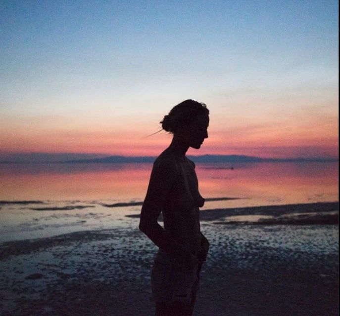 Silhouette of a slender woman standing on a sandy beach at sunset with a pink and blue sky reflecting in the calm water.
