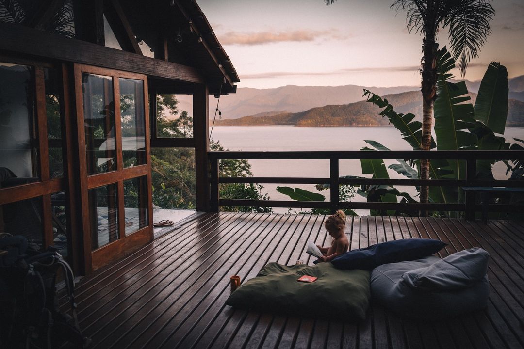 Child reads a book on a wood deck overlooking a serene lake surrounded by lush tropical foliage and distant mountains at dusk.