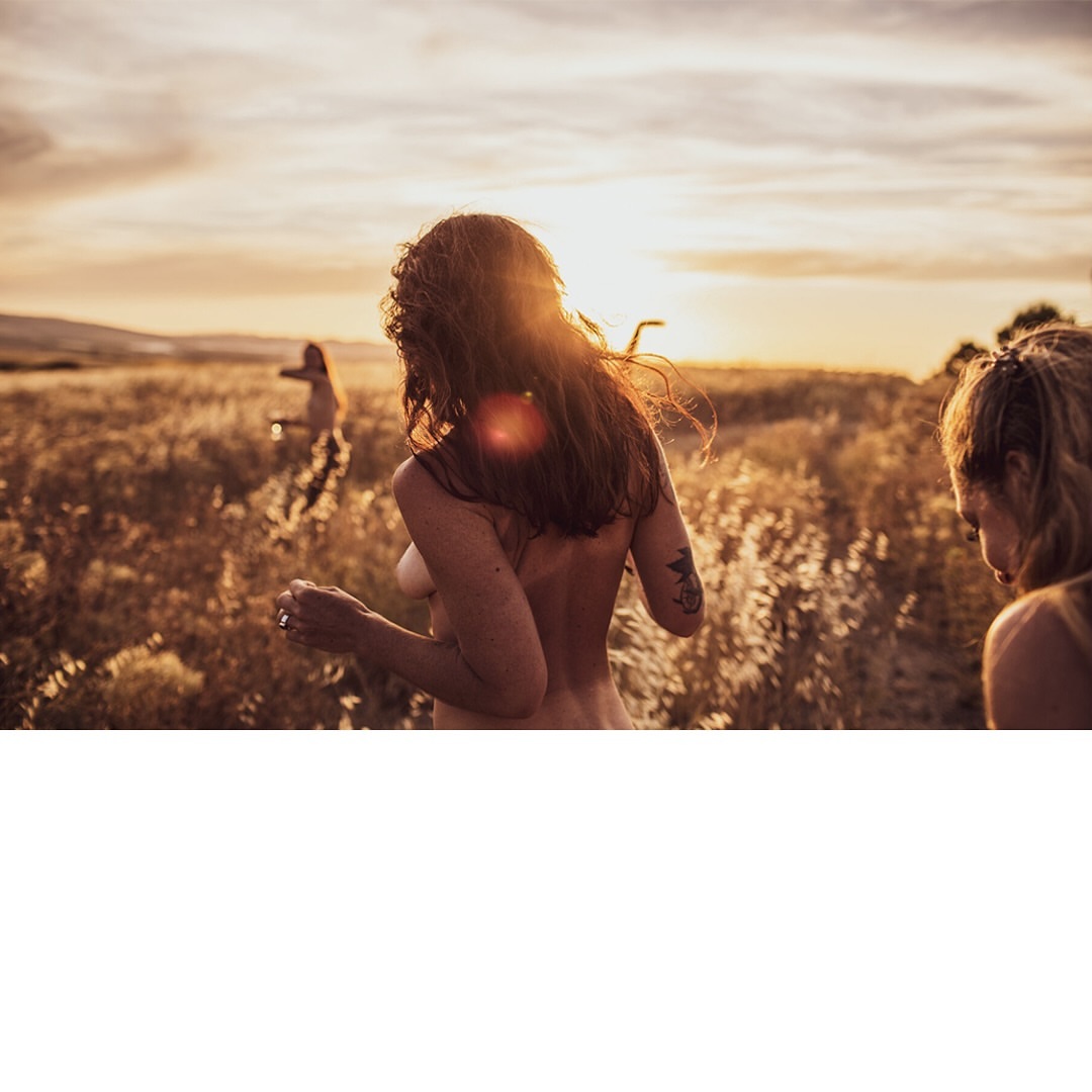 Three topless adult females standing in a field with golden light at sunset, view from behind one of the women.