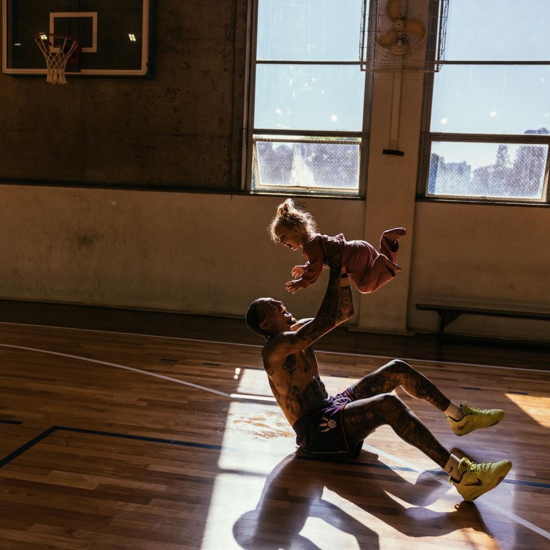 Tattooed man lifts child in the air in a basketball court gym with yellow sneakers, light streaming through windows. 
