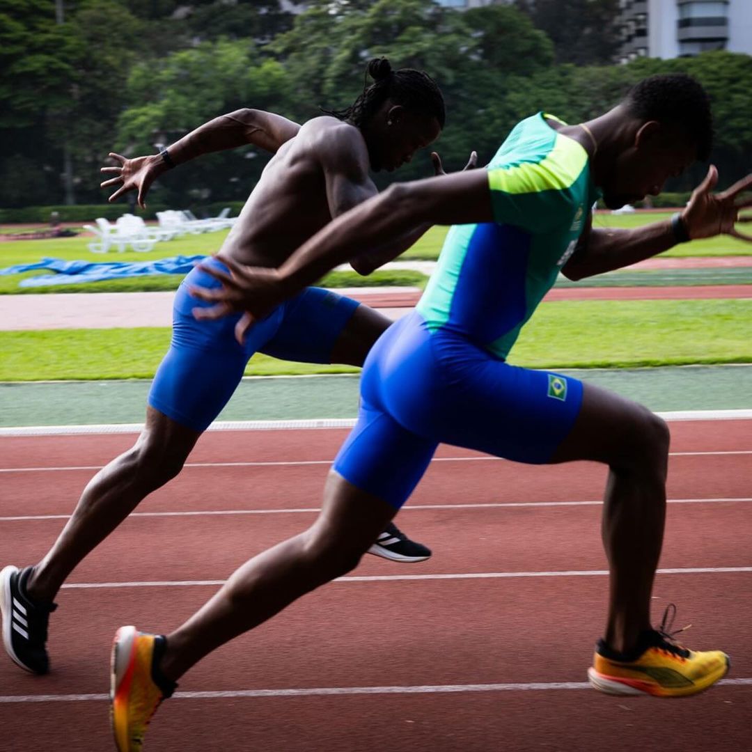 Two athletes sprint on a running track, showcasing their athleticism. One wears a blue uniform with a Brazil flag, the other is shirtless.