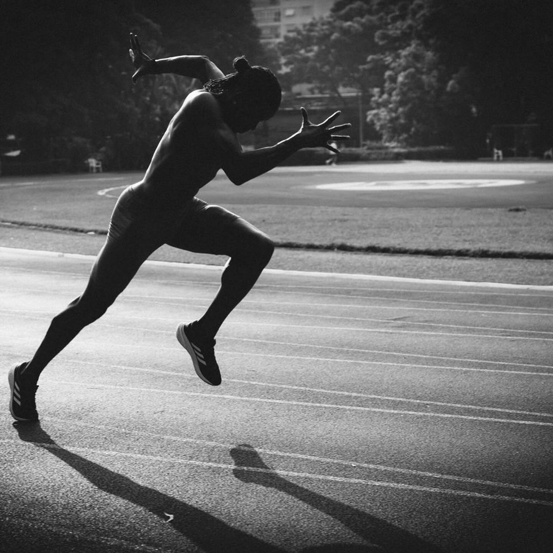 Athlete in starting position on black and white photo. Runner on track preparing to sprint. Trees and building in background.