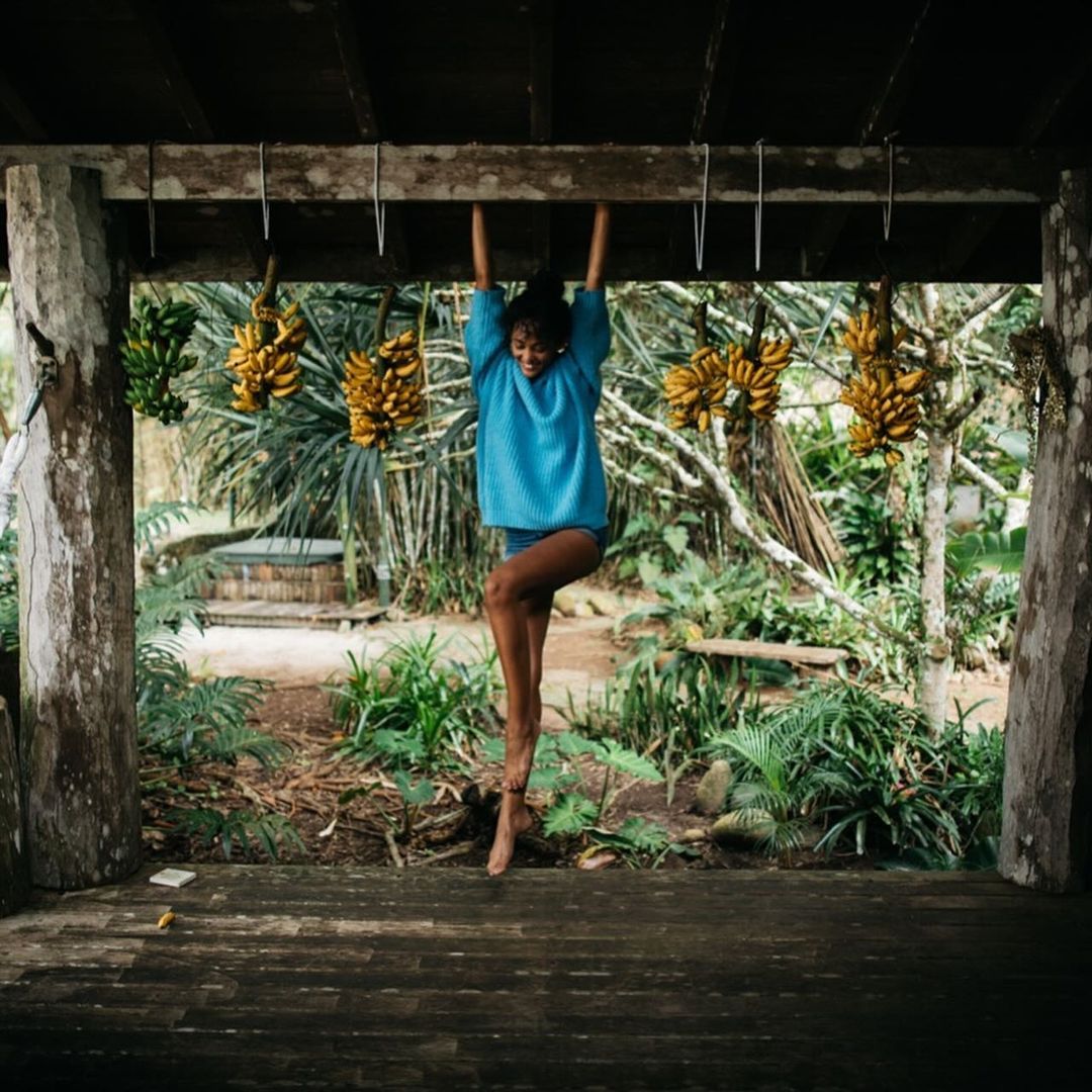 Young woman in blue shirt hangs from wooden beams amidst bananas in a rustic, plant-filled outdoor setting, smiling.